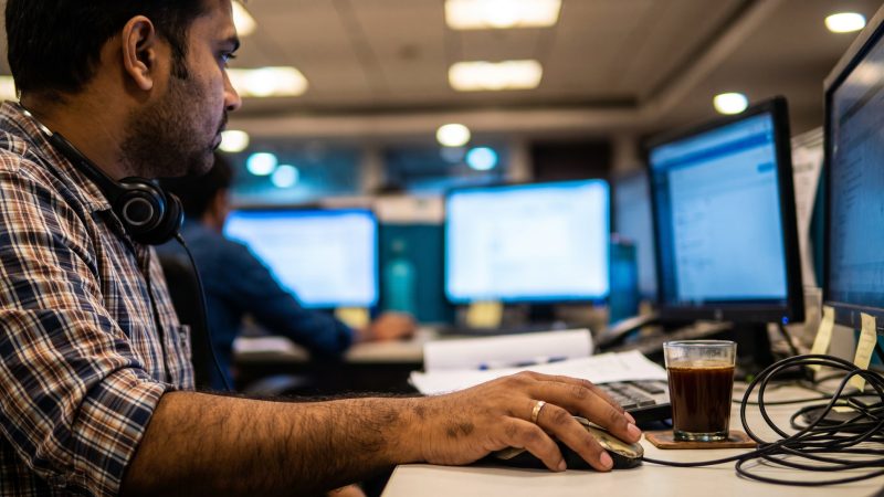 Fraud analyst at a bank operations desk in Bengaluru with monitors out of focus behind his hand on the mouse