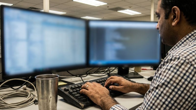 Software developer typing at a worn keyboard with two defocused monitors in an Indian office