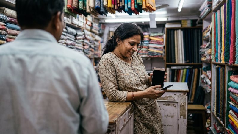Shop owner checking a phone behind the counter of a fabric shop in India while a customer waits